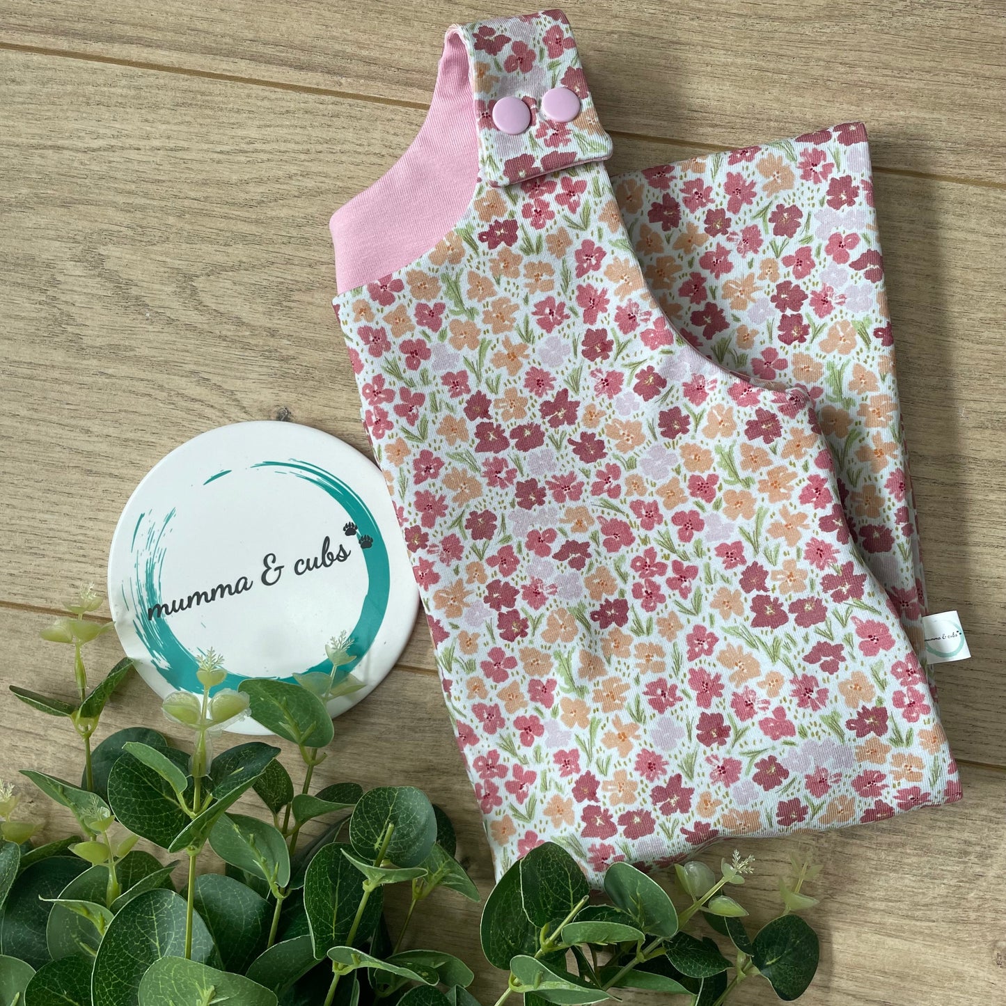 Floral fabric with pink trim on a wooden surface, next to green leaves and a white coaster.