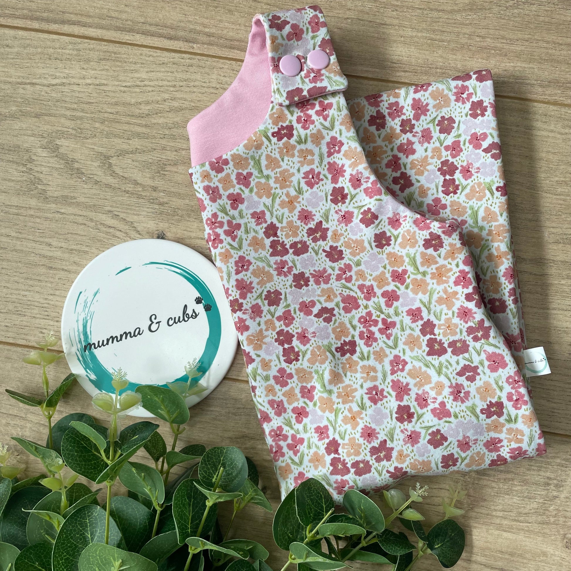 Floral fabric with pink trim on a wooden surface, next to green leaves and a white coaster.