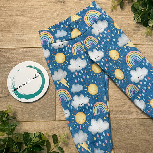 A pair of mid blue leggings with a colorful pattern of rainbows, clouds, and suns, displayed next to a round coaster with 'Mumma & Cubs' branding.